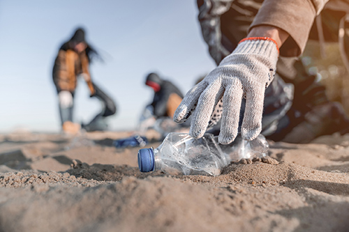 Une main gantée ramasse une bouteille en plastique sur une plage.